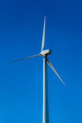 Close up of a white windmill with three blades against clear blue sky. The towering windpump is a generator of alternative and renewable green energy.