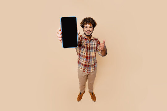 Full body close up top view young Indian man wear brown shirt casual clothes hold in hand use mobile cell phone show thumb up isolated on plain pastel light beige background. People lifestyle concept.
