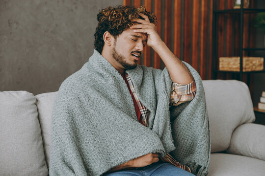 Side view young Indian man wapped in plaid hold head put hand forehead sufer from hangover headache sits on grey sofa couch stay at home hotel flat spend free spare time in living room Lounge concept