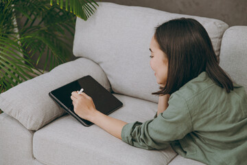 Side view young woman wearing casual clothes graphic designer hold working use write draw stylus pc pen sits on grey sofa couch stay at home hotel flat rest relax spend free spare time in living room.
