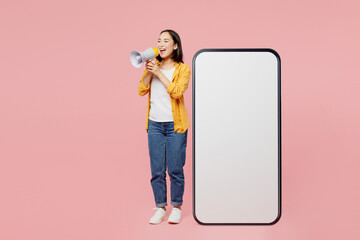 Full body young woman of Asian ethnicity in yellow shirt white t-shirt big huge blank screen mobile cell phone smartphone with area scream in megaphone isolated on plain pastel light pink background