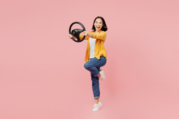 Full body young woman of Asian ethnicity wear yellow shirt white t-shirt hold steering wheel driving car look camera isolated on plain pastel light pink background studio portrait. Lifestyle concept.
