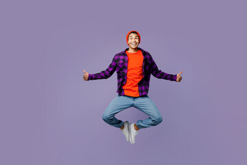 Full body satisfied young man of African American ethnicity wears casual shirt orange hat jump high spread hands show thumb up isolated on plain pastel light purple color background studio portrait.