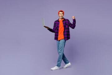 Full body young IT man of African American ethnicity in casual shirt orange hat hold use work on laptop pc computer do winner gesture isolated on plain pastel purple color background studio portrait