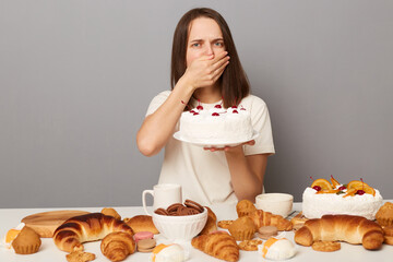 Portrait of sick woman with brown hair wearing white T-shirt sitting at table and feels nausea of sweets, covering mouth with hand, holding cake, isolated over gray background
