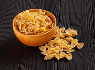 Uncooked farfalle rigate pasta in wooden bowl on black wooden background