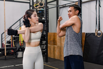 Asian professional muscular male personal trainer teaching female model fit strong body sporty athletic fitness model in sport bra legging standing holding hands arms stretching in CrossFit gym