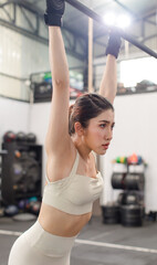 Asian young female fit strong body sporty athletic fitness model in sport bra legging and gloves hanging sweating stretching on metal gymnastic bar take break breathing after exercise training in gym