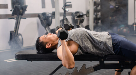 Asian young muscular male fit strong body sporty athletic fitness model in sportswear laying down bench lifting metal dumbbells chest and shoulder training in Crossfit gym with smoke background