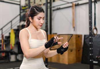 Fototapeta na wymiar Asian young muscular fit strong body sporty athletic sexy female fitness model sitting smiling taking break holding looking at tablet computer mockup after workout exercising weight training in gym