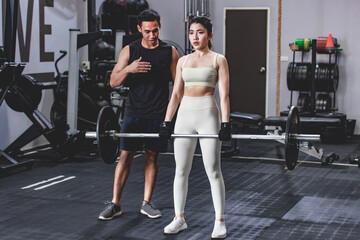 Fototapeta na wymiar Asian professional male personal trainer coaching young muscular fit strong body sporty athletic female fitness in sports bra gloves standing preparing weight trainging with barbell in Crossfit gym