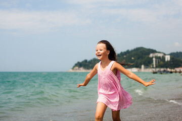 A girl of 6 or 7 with her hair loose and arms outstretched runs along the sea in a pink dress. Middle ground.