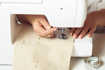 Unrecognizable close up hands of tailor woman using sewing machine, inserting white soft textile pattern with stitch contours under the presser foot. Business production of garments in workshop