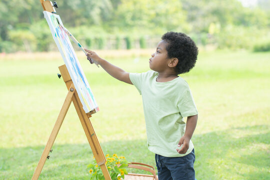 Cute African little boy concentrate with painting and coloring watercolor on canvas in green park on beautiful summer day. Three years old child drawing, outdoor activity. Education in children