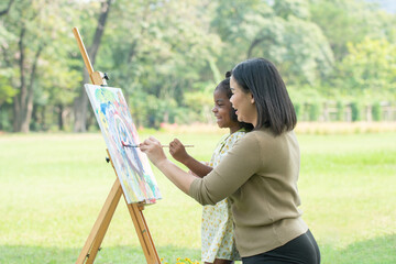Asian mother and cute African daughter smiling holding same paintbrush and painting coloring...