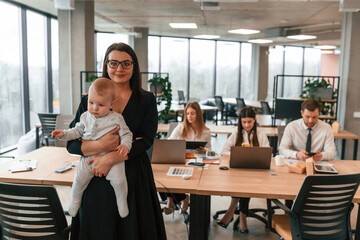 Happy mother is standing and holding her little son. Infant baby is in the office where group of people are working together