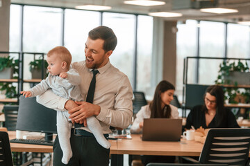 Father is smiling, holding his son. Infant baby is in the office where group of people are working together