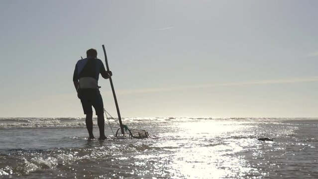 Back view of man in wetsuit collecting shellfish in grill basket on the shore of the beach at sunset