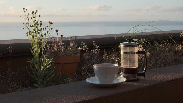 Caf&eacute; caliente humeante de cafetera de &eacute;mbolo sobre el muro de una jardinera con luz de amanecer en una terraza con vistas al mar y mano que pone la taza en el plato