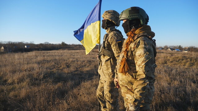 Military couple of ukrainian army lifted blue-yellow banner holding hands of each other at field. Young soldiers in camouflage uniform standing with a waving Ukraine flag at meadow. Victory at war