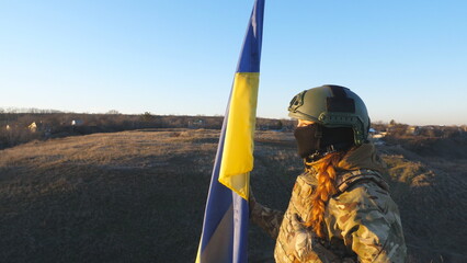 Young female soldier in camouflage uniform lifting flag of Ukraine. Ukrainian army woman stands to...