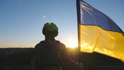 Ukrainian army man stands with lifted national banner against background of sunset. Young male soldier in military uniform and helmet holds a waving flag of Ukraine to peak of hill. End of war concept