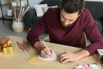 Man writing message in greeting card at wooden table in room