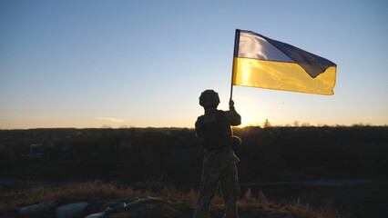 Military man in uniform waving flag of Ukraine against background of sunset. Male ukrainian army soldier lifting national banner at hill in honor of the victory against russian aggression. Rear view