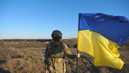 Dolly shot of male soldier in military uniform and helmet holds a waving flag of Ukraine. Ukrainian army man stands with lifting national banner at field. Victory against russian aggression. Slow mo