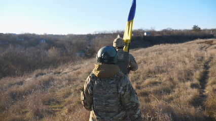 Female and male soldier of ukrainian army walking with flag of Ukraine at field. Woman and man in military uniform going with national banner at meadow as sign of victory against russian aggression