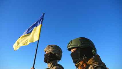 Young soldiers of ukrainian army standing at peak of hill with raised national banner. Woman and man in camouflage uniform looking at distance with lifted flag of Ukraine against blue sky. Close up