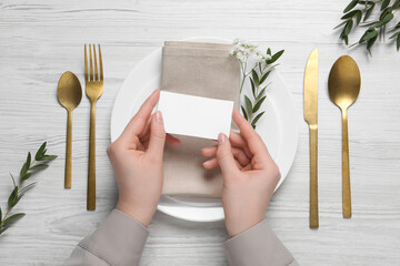 Woman holding blank card near stylish setting at white wooden table, top view. Space for text