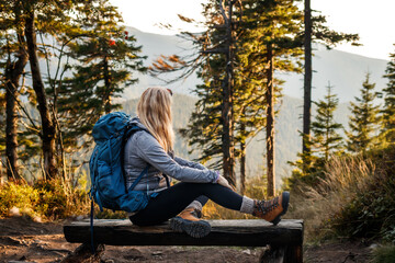 Relaxation during hiking in mountain. Woman with backpack resting on wooden bench in forest....
