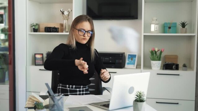 The young woman in glasses is sitting at her workstation in front of a laptop. She is discontentedly shuffling through a few sheets of paper, expressing dissatisfaction and frustration with her work