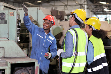 Technician engineer in protective uniform with hardhat standing and teaching apprentices or colleague worker to use computerized machine control at heavy industry manufacturing factory