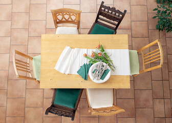 Preparation for dinner with table runner, white plates, cutlery, green napkins and some tulip flowers, six different wooden chairs, tiled terracotta floor, top view from above