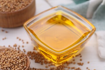 Bowl of natural oil and mustard seeds on white table, closeup