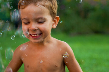 A little boy without clothes happily stands in a stream of flying soap bubblesA little boy without...