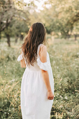 Fototapeta premium Rear view of young woman in summer white dress walking in the park. Attractive brunette girl looking into the distance at nature