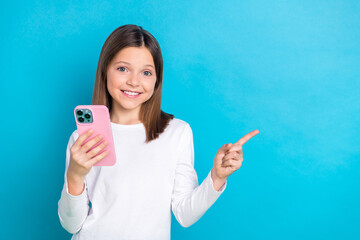 Photo of positive schoolgirl straight hairdo dressed white shirt hold smartphone directing empty space isolated on blue color background