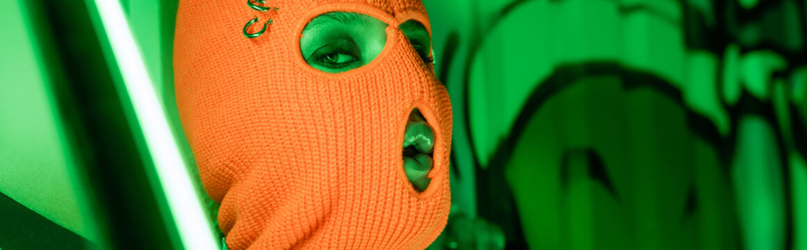 portrait of sexy woman in knitted orange balaclava near bright neon lamp and green wall with graffiti, banner.