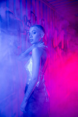 seductive short haired woman in silver top looking at camera near wall with graffiti in blue and pink light with smoke.