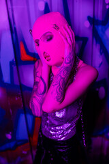 passionate tattooed woman touching pink balaclava while standing with closed eyes near colorful graffiti in purple light.