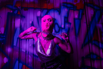 Fototapeta na wymiar passionate tattooed woman in pink balaclava and metallic top posing with chain near wall with graffiti in purple lighting.