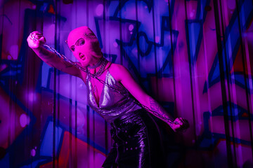 seductive woman in metallic top and balaclava holding silver chain near wall with colorful graffiti in purple light.
