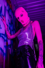 low angle view of provocative tattooed woman in shiny top and pink balaclava posing near graffiti in purple neon light.