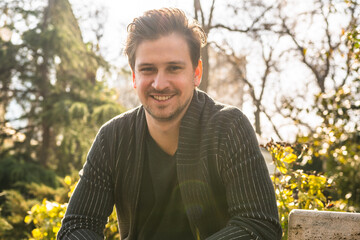 A man sits in a park in the sunlight wearing a black jacket with a black shirt