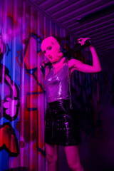 stylish woman in balaclava and metallic top posing with baseball bat near wall with graffiti in purple neon light.