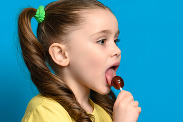 On a blue background, a girl licks a round lollipop close-up - obrazy, fototapety, plakaty