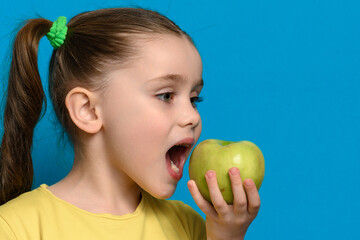 A little girl is holding and biting a green apple on a blue background.